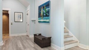A hallway with a wooden floor, a wicker basket, a painting of sailboats, and a door leading to another room. - Ocean Views, Elevator, Fire Pit (Surf City)