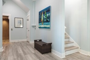 A hallway with a wooden floor, a wicker basket, a painting of sailboats, and a door leading to another room. - Ocean Views, Elevator, Fire Pit (Surf City)