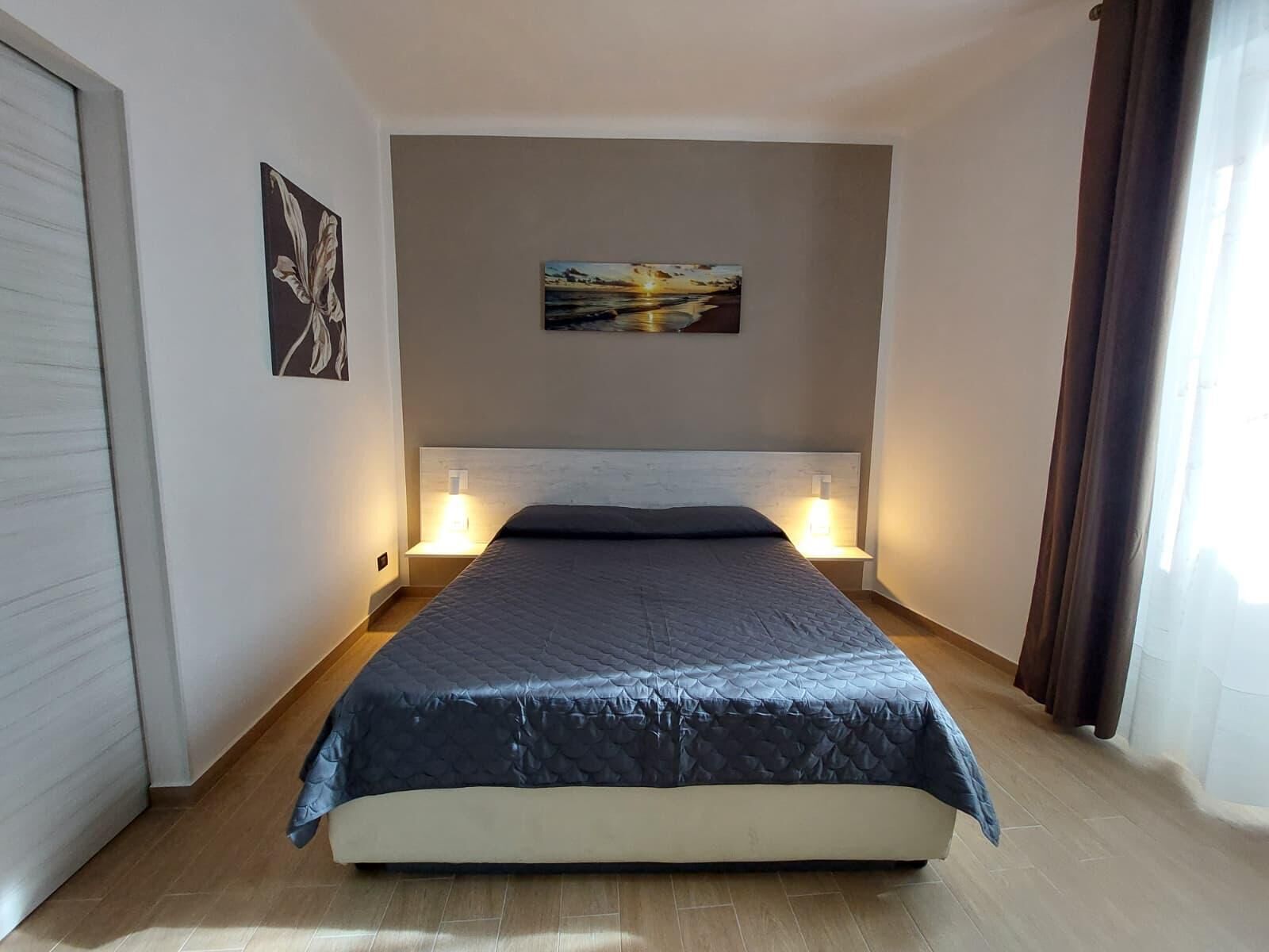 A bedroom with a bed, two bedside lamps, a painting on the wall, and a window with curtains.