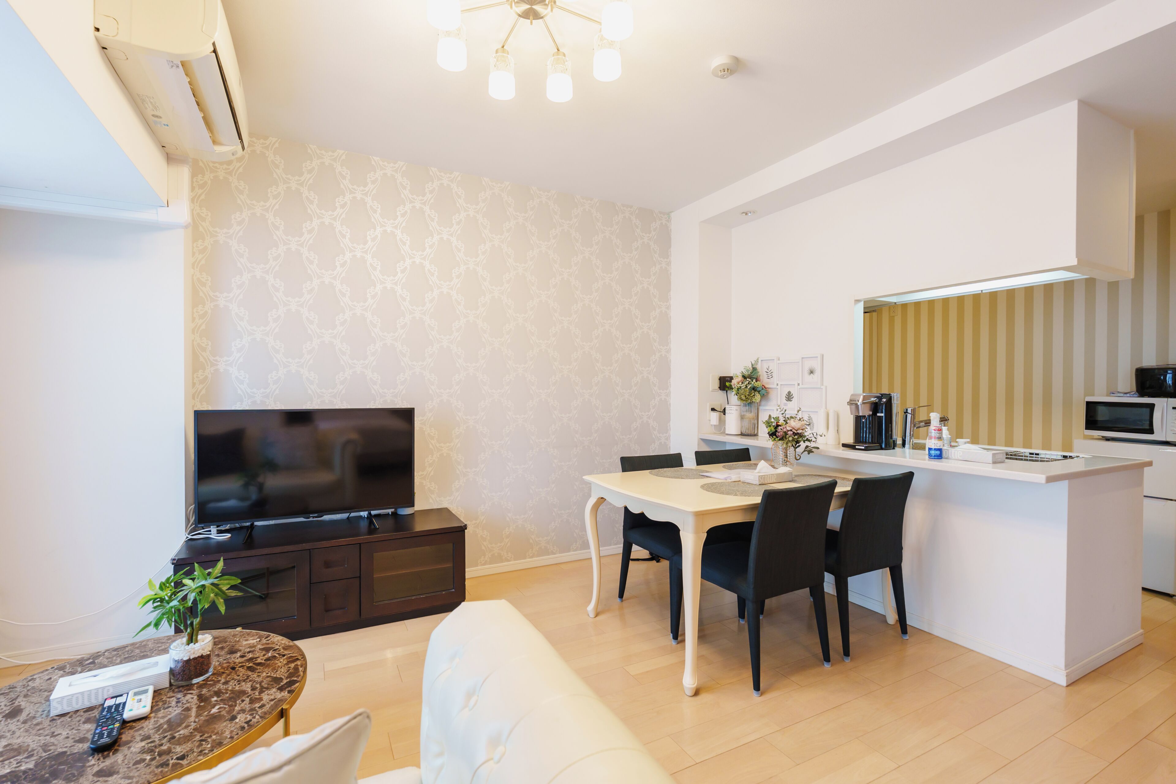 A modern living room with a flat-screen TV, a dining area with a white table and dark chairs, and a kitchen with a white countertop and appliances.