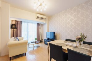 A modern living room with a sofa, a dining table with chairs, a television, and an air conditioning unit. - SERENE | Kuromon · 1006 | 60 m² · 2BR | Sleeps 6 | Family Suite | 2 min to Kuromon (大阪市)