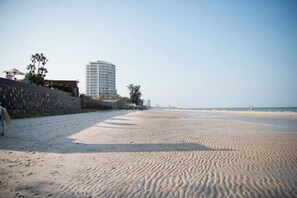 Ein sandiger Strand mit einer Steinmauer, Palmen und einem Hochhaus im Hintergrund.