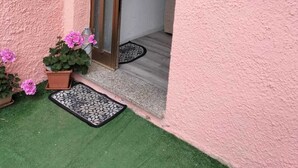 Paret exterior rosa amb una porta que condueix a una habitació. Hi ha dues plantes en testos amb flors roses al sòl.