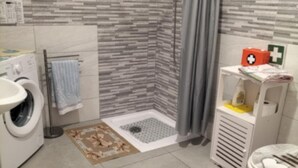 Un bany amb rentadora, una dutxa amb cortina gris, un vàter i una petita estant amb un kit de primers auxilis.