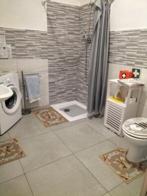A bathroom with a washing machine, a shower with a grey curtain, a toilet, and a small shelf with a first aid kit. - Vacation home in Santa Caterina di Pittinuri, a hamlet of Cuglieri (Or), near the beach (Santa Caterina di Pittinuri)