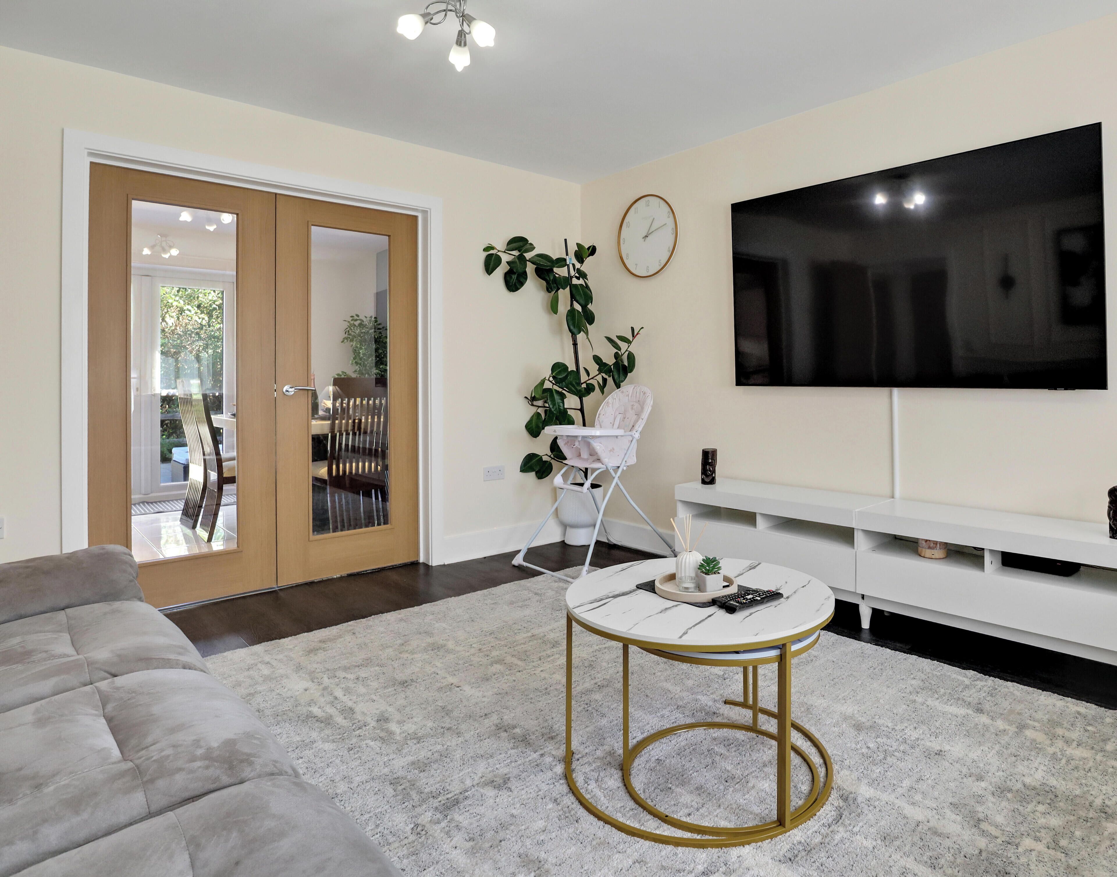 A modern living room with a grey sofa, a round coffee table with gold legs, a white TV stand, and a large flat-screen TV.