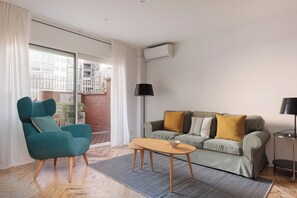 Ein modernes Wohnzimmer mit einem petrolfarbenen Sessel, einem grauen Sofa mit Senfgelb-Kissen, einem Holztisch und einer schwarzen Stehlampe.