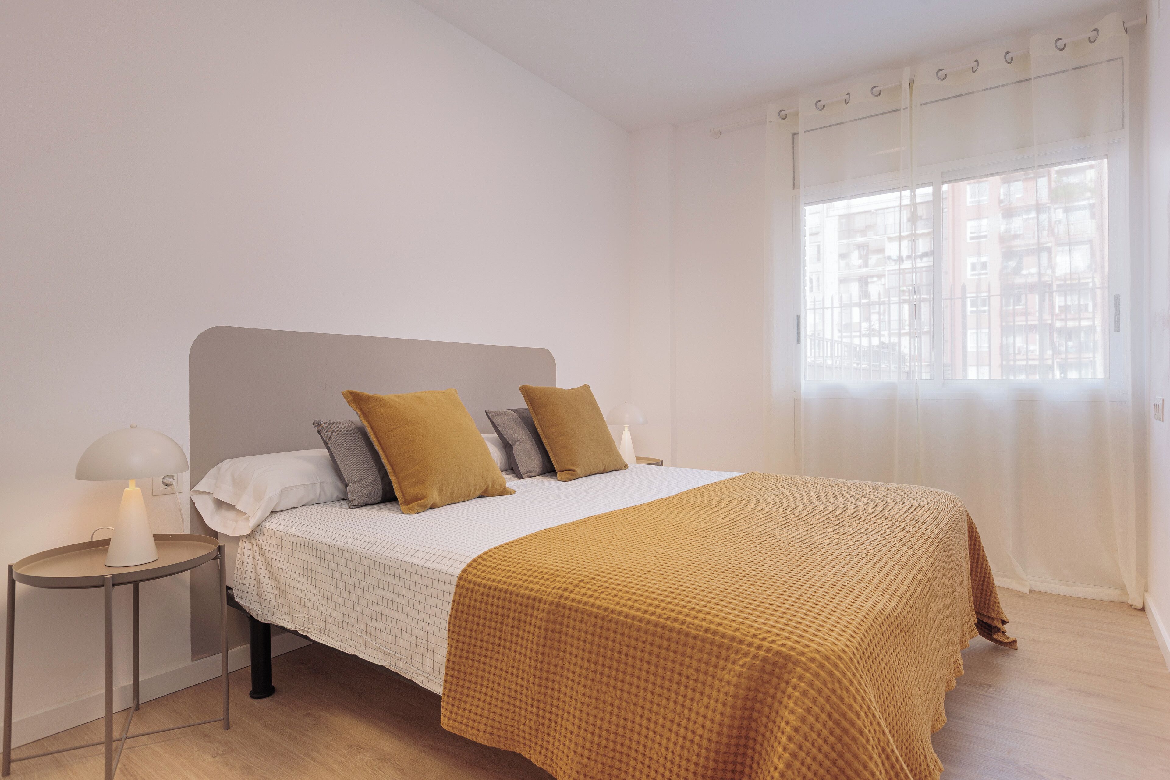 Ein ordentlich bezogenes Bett mit weißer und brauner Decke, eine Nachttischlampe und Blick auf ein Gebäude durch ein Fenster.