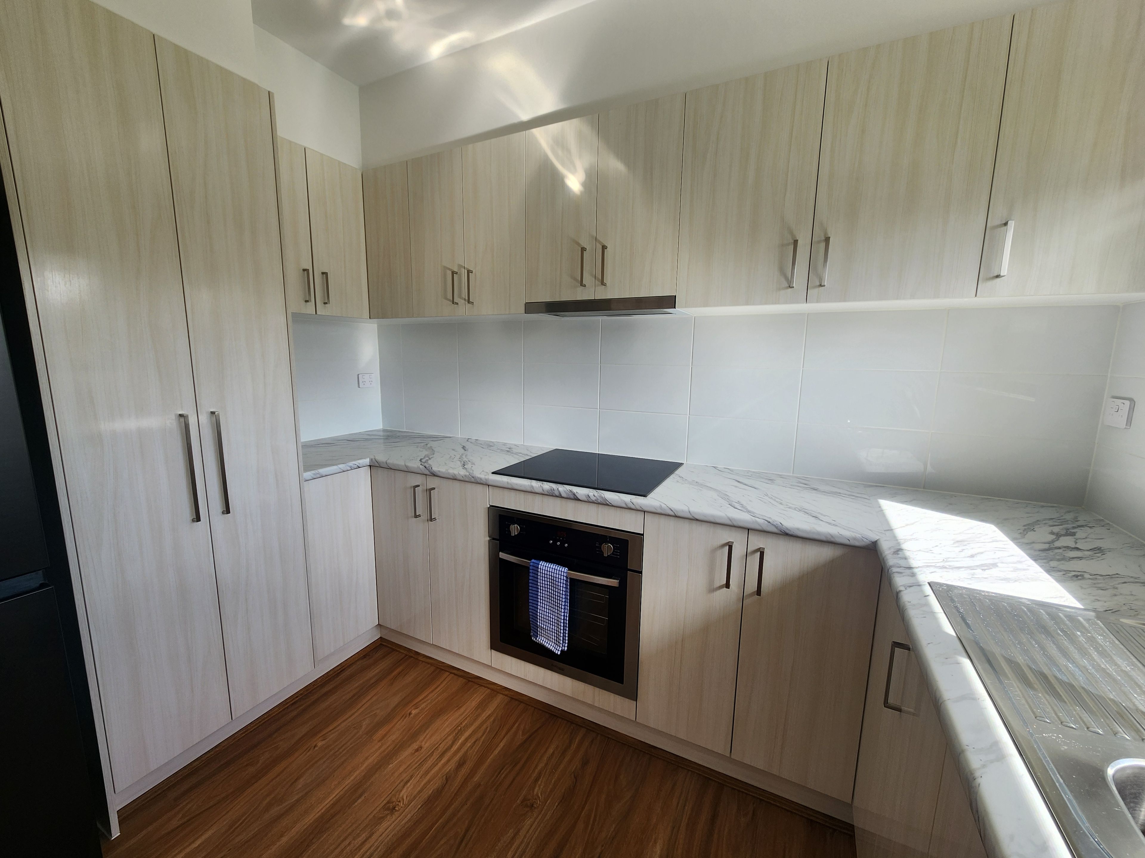 A modern kitchen with light wood cabinets, a built-in oven, and a marble countertop.