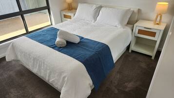 Una cama bien hecha con una manta azul y una toalla enrollada, dos mesitas de noche con lámparas, y una ventana con vistas al exterior.
