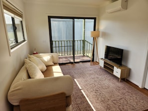 A living room with a sofa, a television on a stand, a floor lamp, and a view through a glass door to an outdoor balcony.