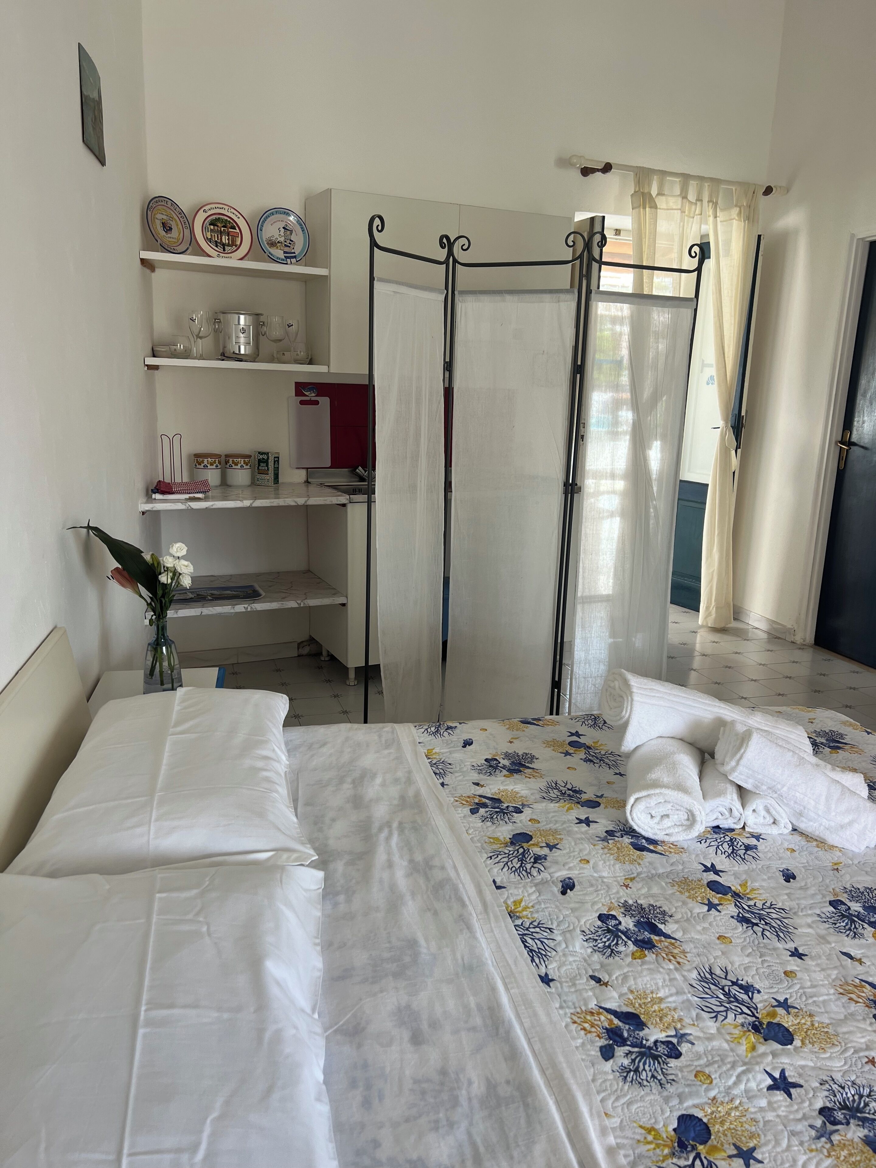 A hotel room with a bed, a bedside table, a vase with flowers, a wardrobe, a shelf with decorative plates, and a curtain.
