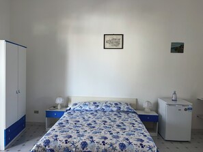 A bedroom with a bed, a wardrobe, a nightstand, a bedside table, a lamp, and a refrigerator.