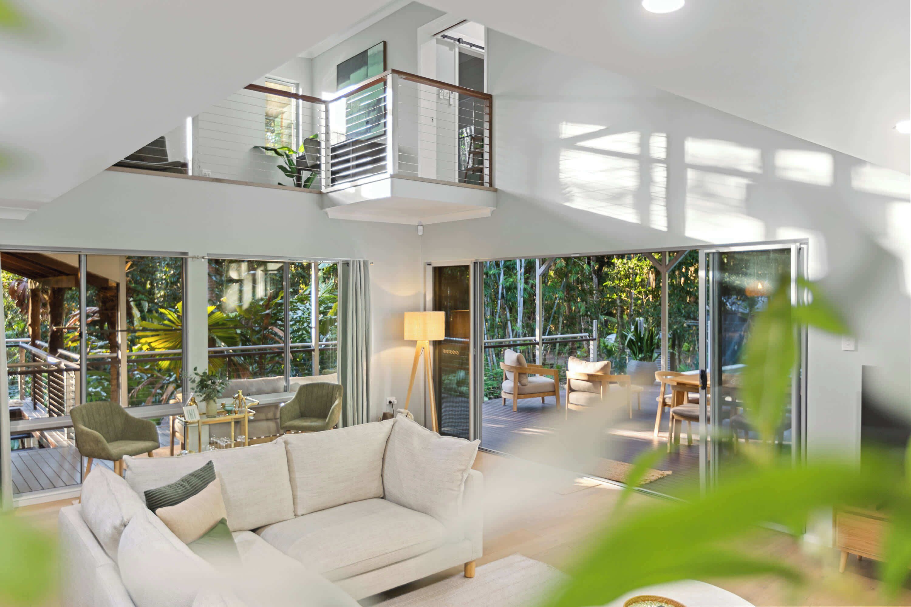 A spacious living area with a high ceiling, large windows, and a balcony overlooking greenery.