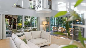 A modern living room with a sectional sofa, a round coffee table, and large glass doors leading to an outdoor area with a view of greenery. - Private Kuranda Rainforest Villa with Pool (Kuranda)