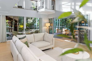 Ein modernes Wohnzimmer mit einer Eckcouch, einem runden Couchtisch und großen Glasfenstern, die zu einem Außenbereich mit Blick auf Grünflächen führen.