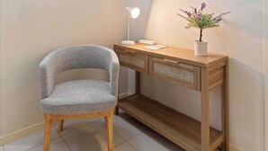 Uma cadeira com encosto trançado, uma mesa consola de madeira com uma luminária e um vaso de planta, e um chão em mosaico.