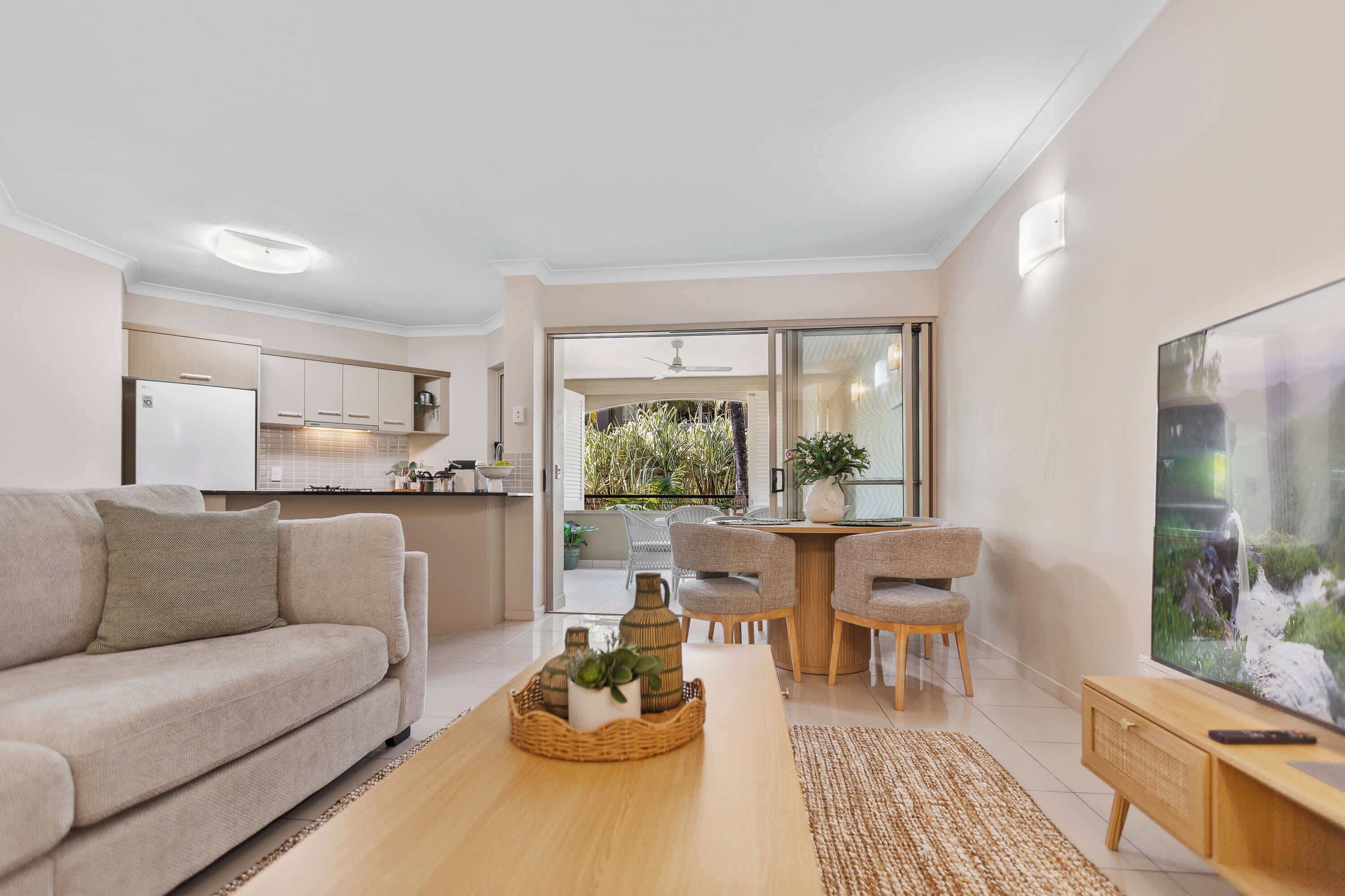 A modern living room with a sofa, a coffee table, and a TV. There is a kitchen area with cabinets and a refrigerator. A dining area with chairs and a table is visible through an open door.