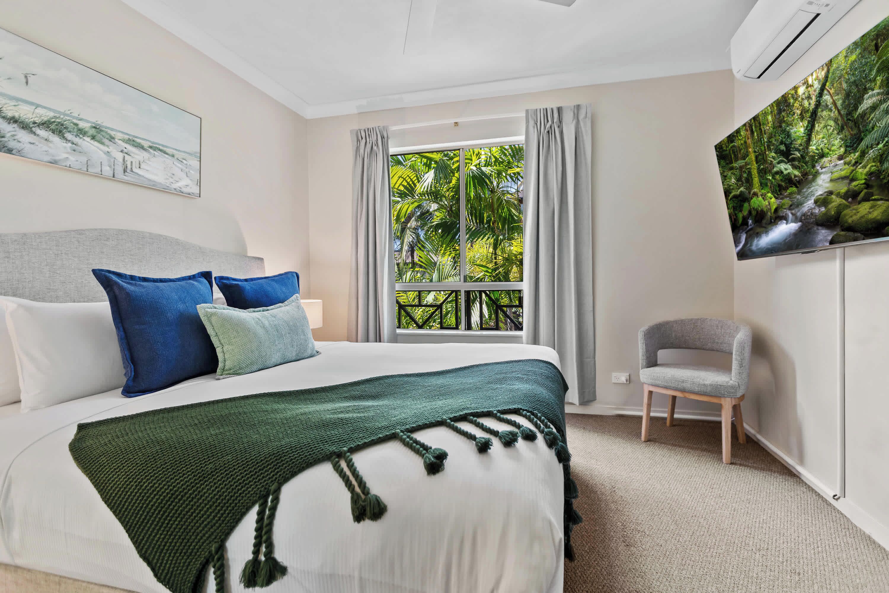 A hotel room with a bed, a chair, a window with a view of greenery, and an air conditioning unit.