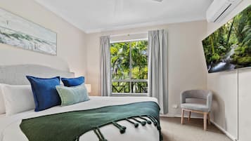 A hotel room with a bed, a chair, a window with a view of greenery, and an air conditioning unit.