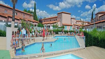 A swimming pool area with multiple pools, surrounded by a fence, and buildings with red-tiled roofs in the background.