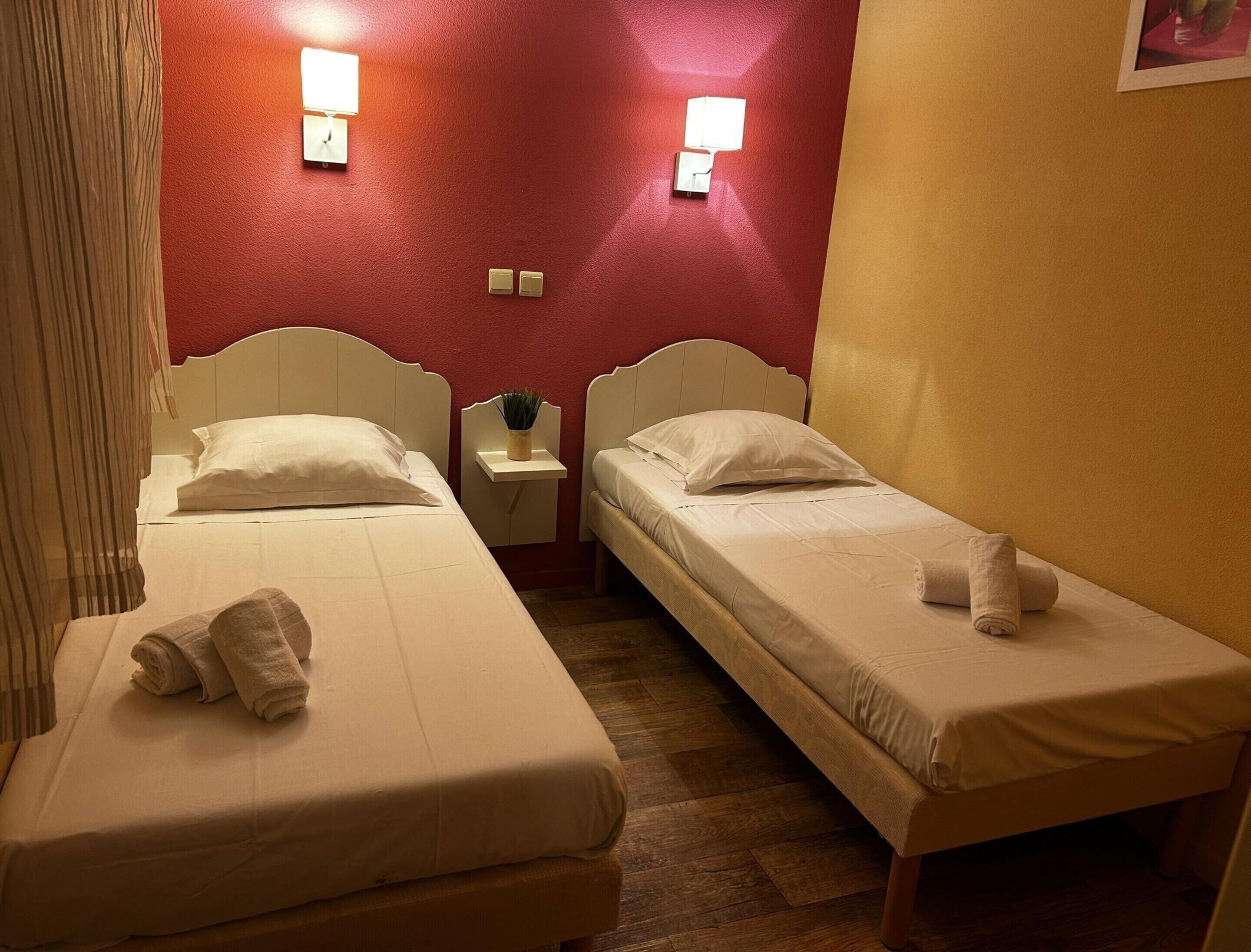 Two single beds with white linens, a small bedside table with a lamp, and a wall-mounted light fixture.