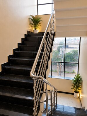 Una escalera con escalones negros y barandilla metálica, que lleva a un rellano con una planta en maceta.