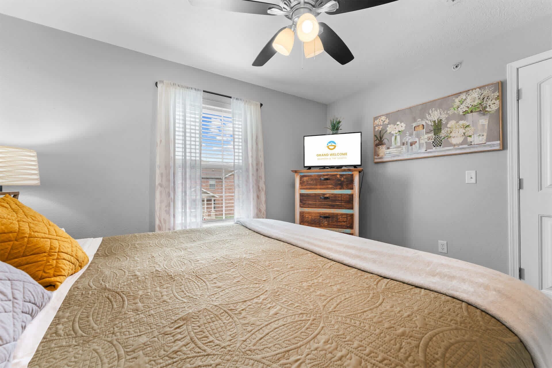 A bedroom with a bed, a ceiling fan, a TV on a wooden stand, and a painting on the wall.