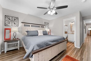 Ein Schlafzimmer mit Bett, Nachttisch, Lampe, Deckenventilator, Kunstwerk und Blick ins Badezimmer.