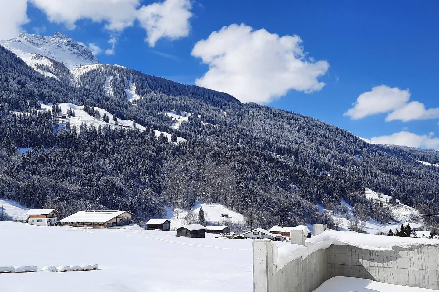 A snowy mountain landscape with a clear sky, a few buildings, and a wooden fence.