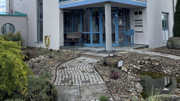 Un bâtiment moderne doté de grandes fenêtres, d’une entrée abritée et d’un chemin de pierre menant à un élément décoratif aquatique.