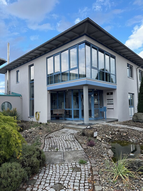 A modern building with large windows, a covered entrance, and a stone pathway leading to a water feature.