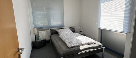 A bedroom with a bed, bedside tables, a window with blinds, a radiator, and a ceiling light fixture.