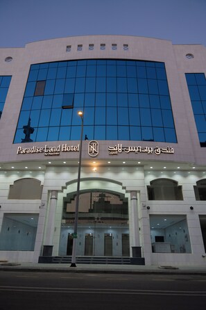 The exterior of the Paradise Land Hotel with a modern architectural design, featuring a prominent entrance and a large glass facade reflecting the sky.