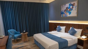 A hotel room with a large bed, a desk, a chair, and a painting on the wall.