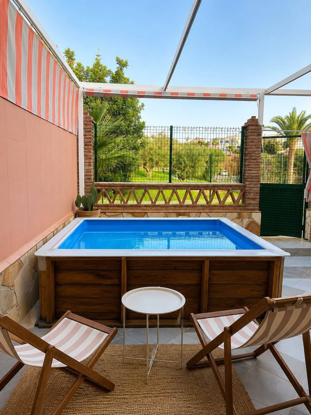 A swimming pool with wooden chairs and a small table, surrounded by a covered patio area with a view of a green landscape.