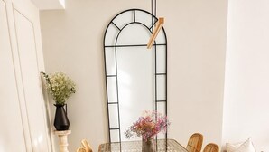 Ein Essbereich mit einem Glastisch, umgeben von Korbstühlen, einem schwarzen Metallbogen und einem Topfpflanzen.