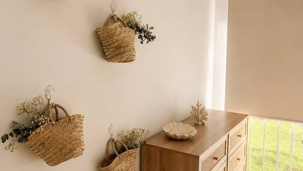Ein Zimmer mit einem Holzkasten, an der Wand befestigten gewebten Körben mit Grünpflanzen und Blick ins Freie durch ein Fenster.