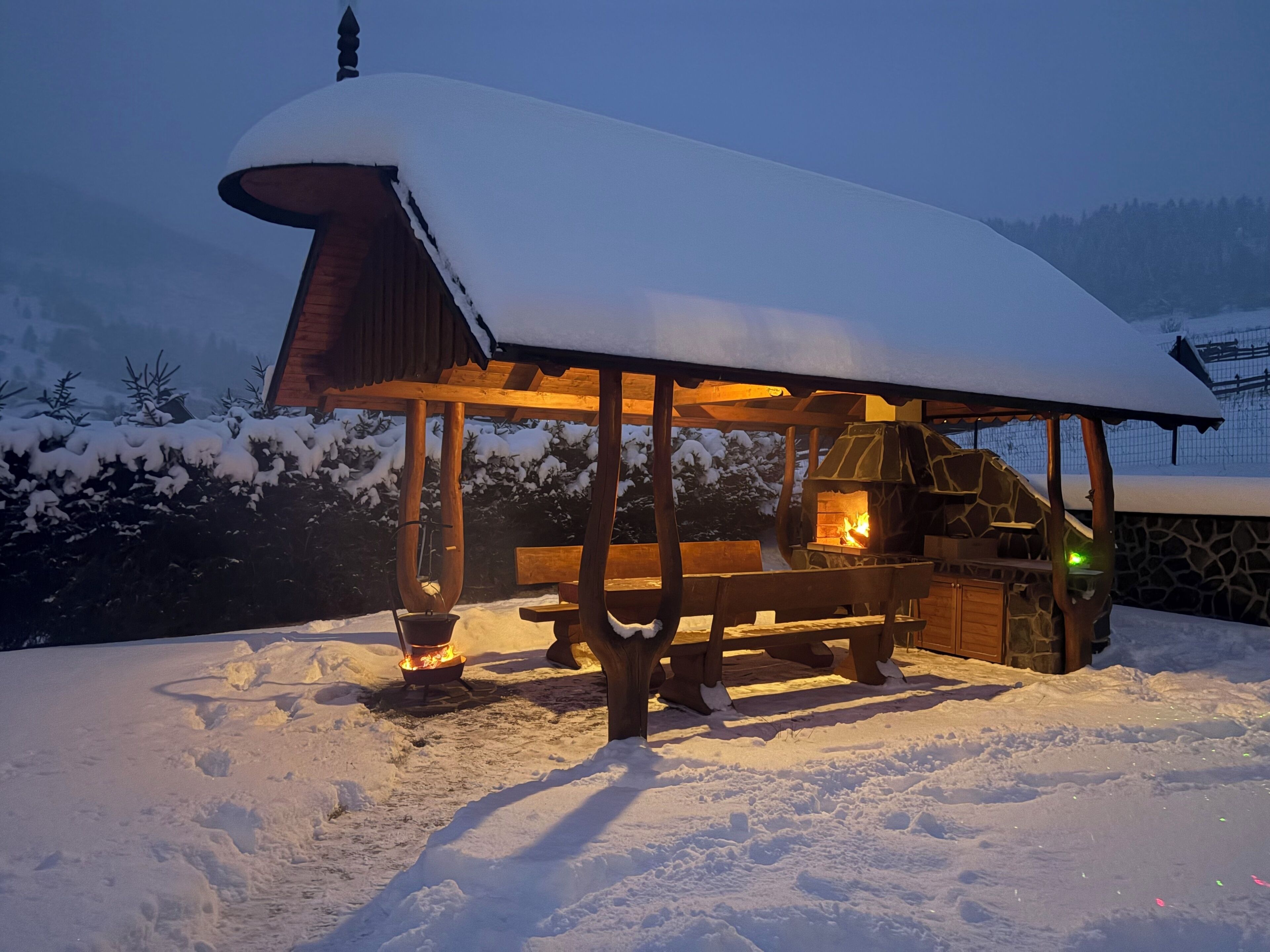 Eine hölzerne Außenkonstruktion mit Steinofen, Bänken und Feuerstelle in verschneiter Landschaft.