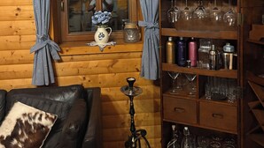 Ein gemütliches Zimmer mit Holzvertäfelung, einem Fenster mit Vorhängen, einem Bücherregal mit diversen Gegenständen und einer Wasserpfeife auf dem Boden.