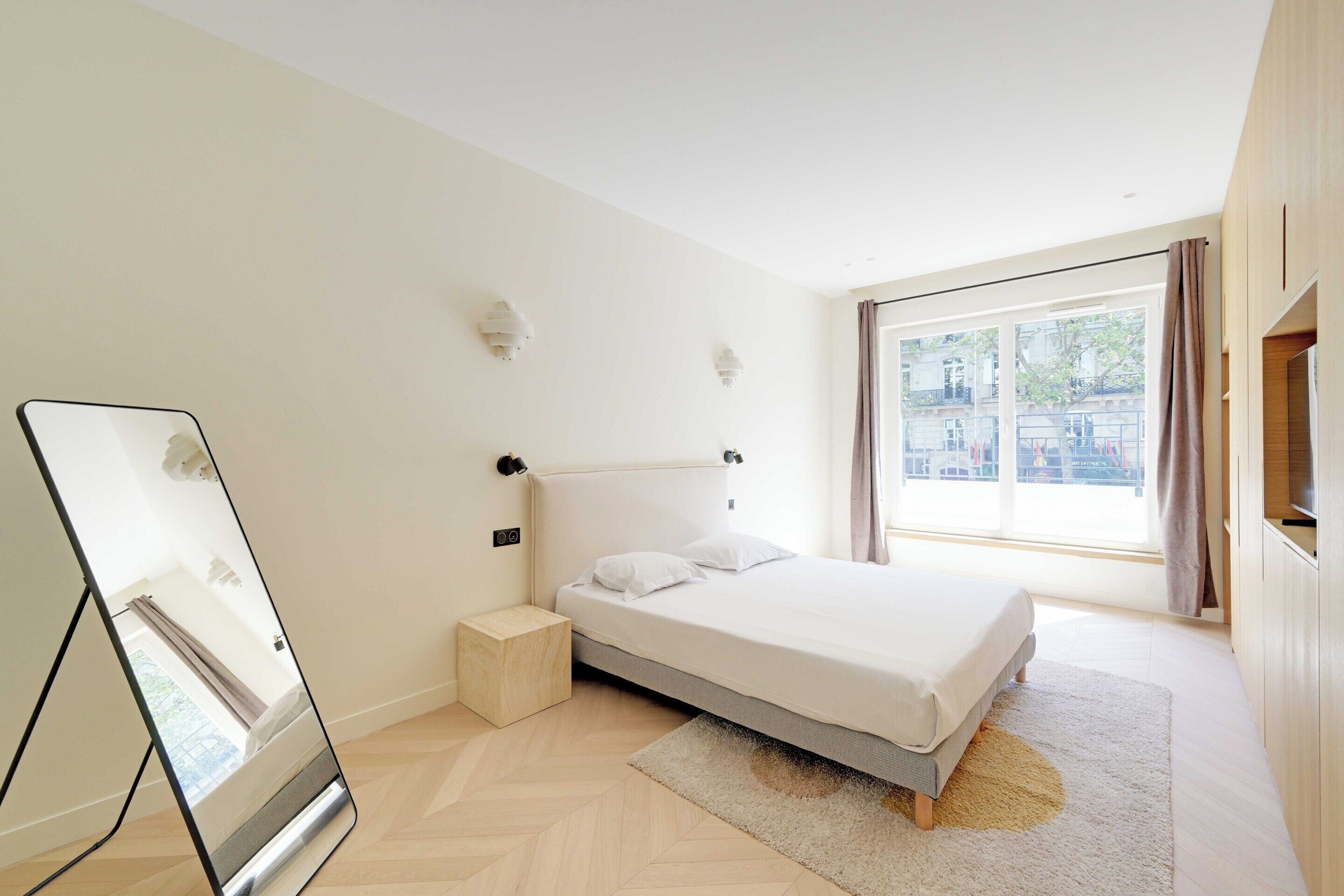 Een slaapkamer met een bed, nachtkastje, spiegel, raam met gordijnen en een televisie.