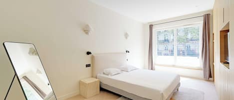 A bedroom with a bed, a bedside table, a mirror, a window with curtains, and a television.