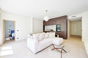 A modern living room with a white sectional sofa, a glass coffee table, and a kitchen area with dark wood cabinetry and a white countertop. - Modern a/c - 2bdr/4p Flat - Montorgueil (Paris)