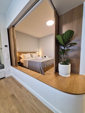 Ein modernes Hotelzimmer mit großem Fenster, einem Bett, einem Nachttisch, einer Zimmerpflanze und einer eingebauten Sitzbank.