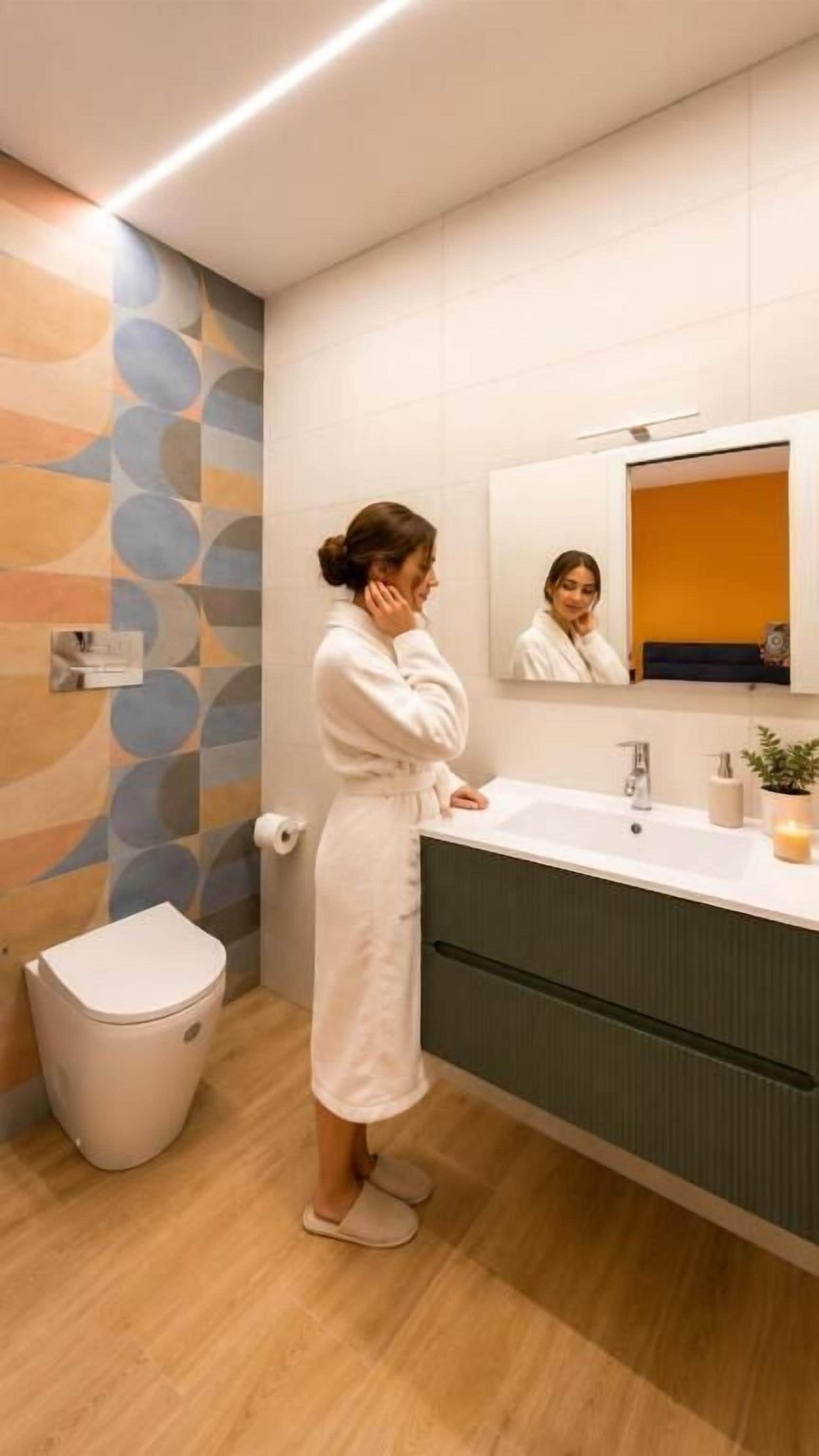 A person in a white bathrobe standing in a modern bathroom with a sink, mirror, and a trash bin.