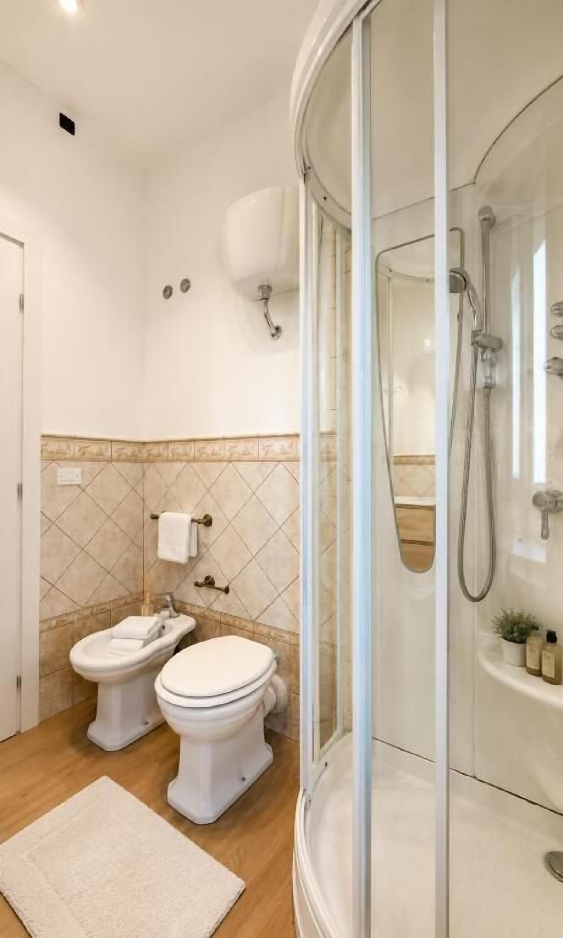 A bathroom with a glass-enclosed shower, two toilets, a sink, and a small plant.