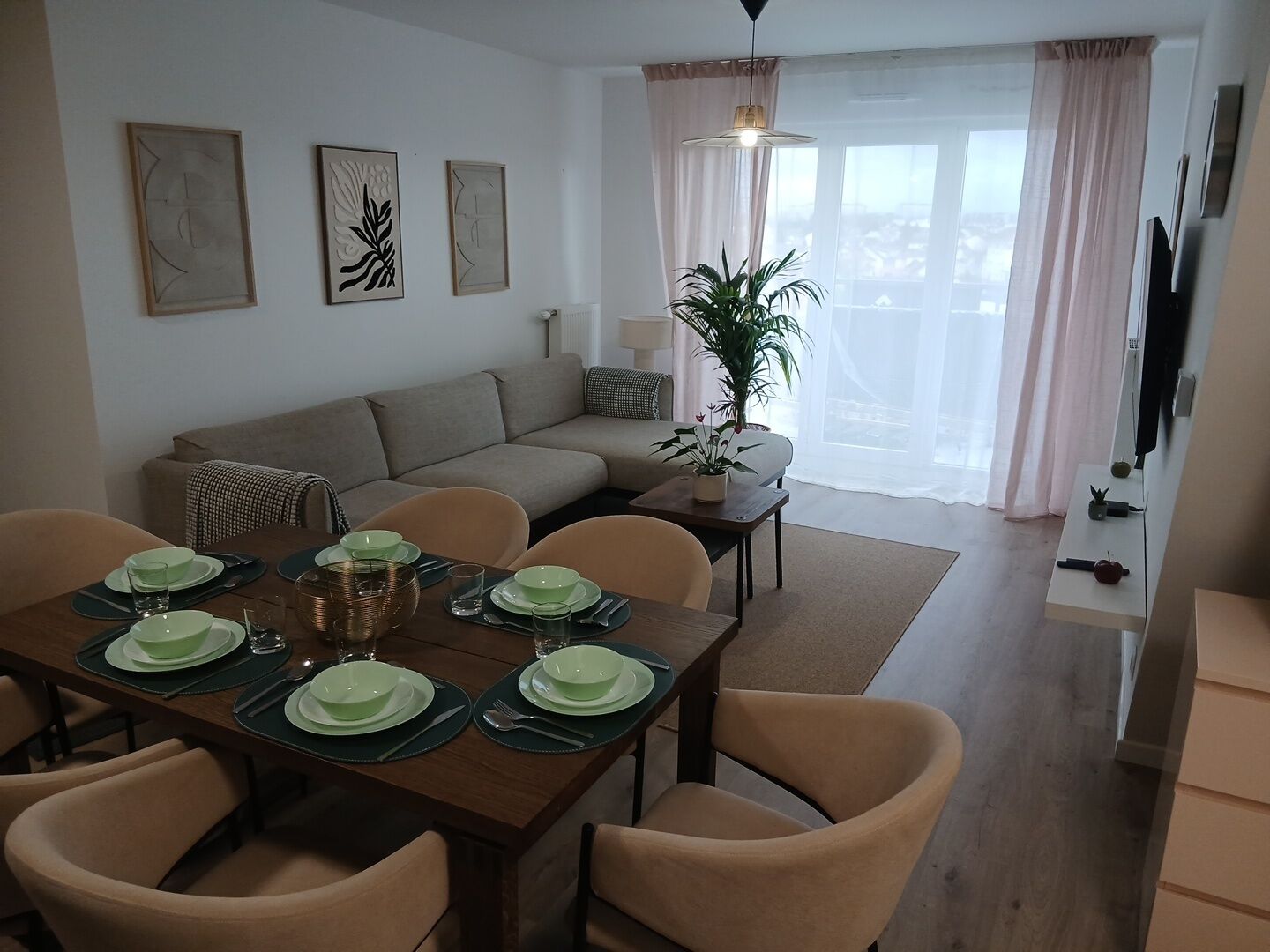 A dining area with a table set for four, a sofa, a plant, and a TV mounted on the wall.
