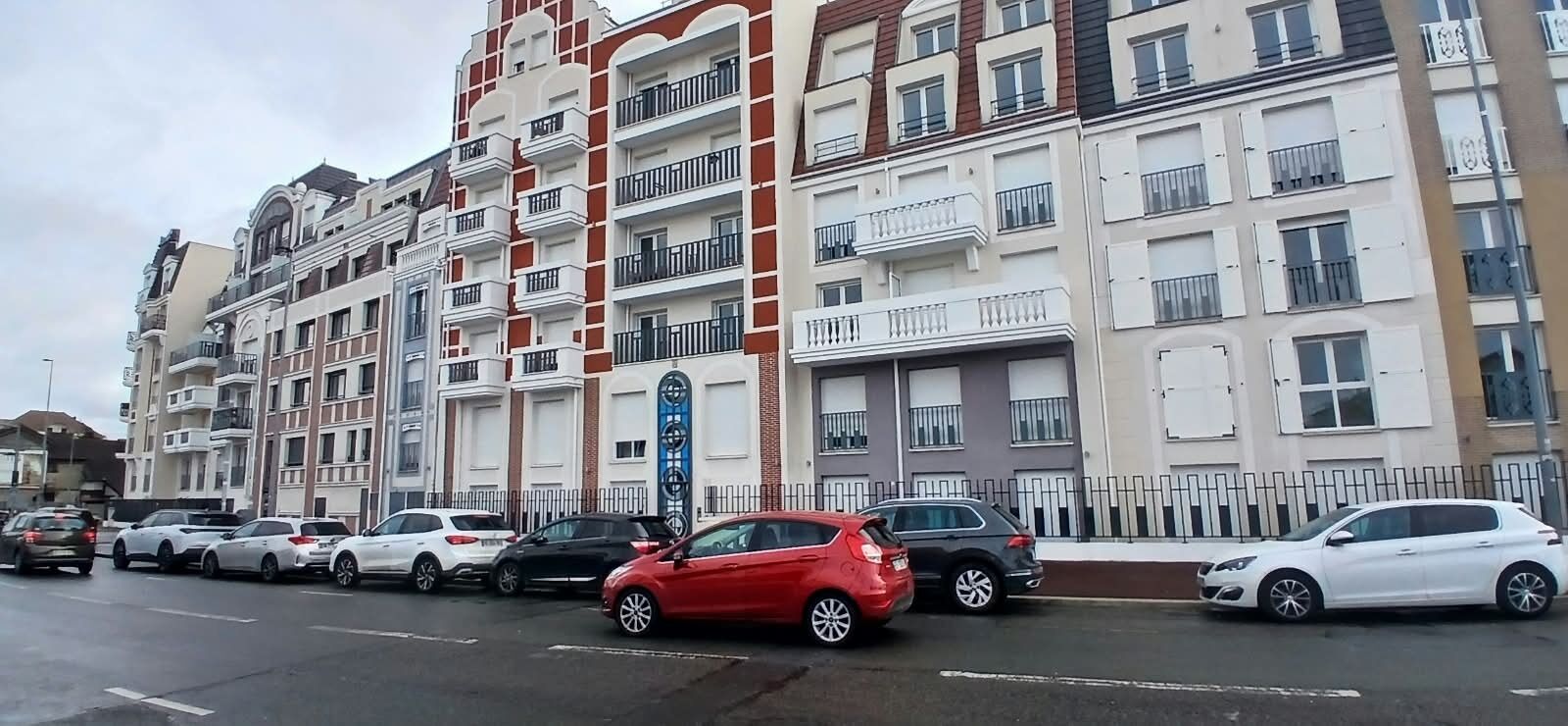 A multi-story residential building with balconies, parked cars, and a clear sky.