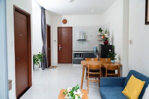 A modern living room with a sofa, a dining table with chairs, a kitchen area with a stove and microwave, and a hallway with a door and a potted plant. - Green Apartment near My Khe Beach and Dragon Bridge- 201 (Đà Nẵng)