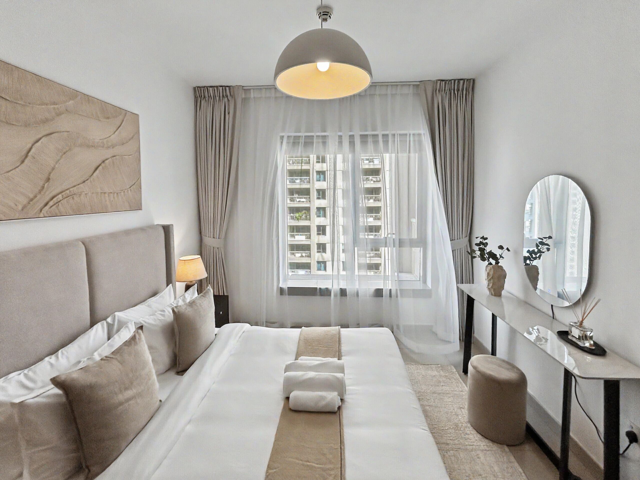 Une chambre moderne avec un grand lit, une table de chevet, un miroir et une vue sur la ville à travers des rideaux transparents.